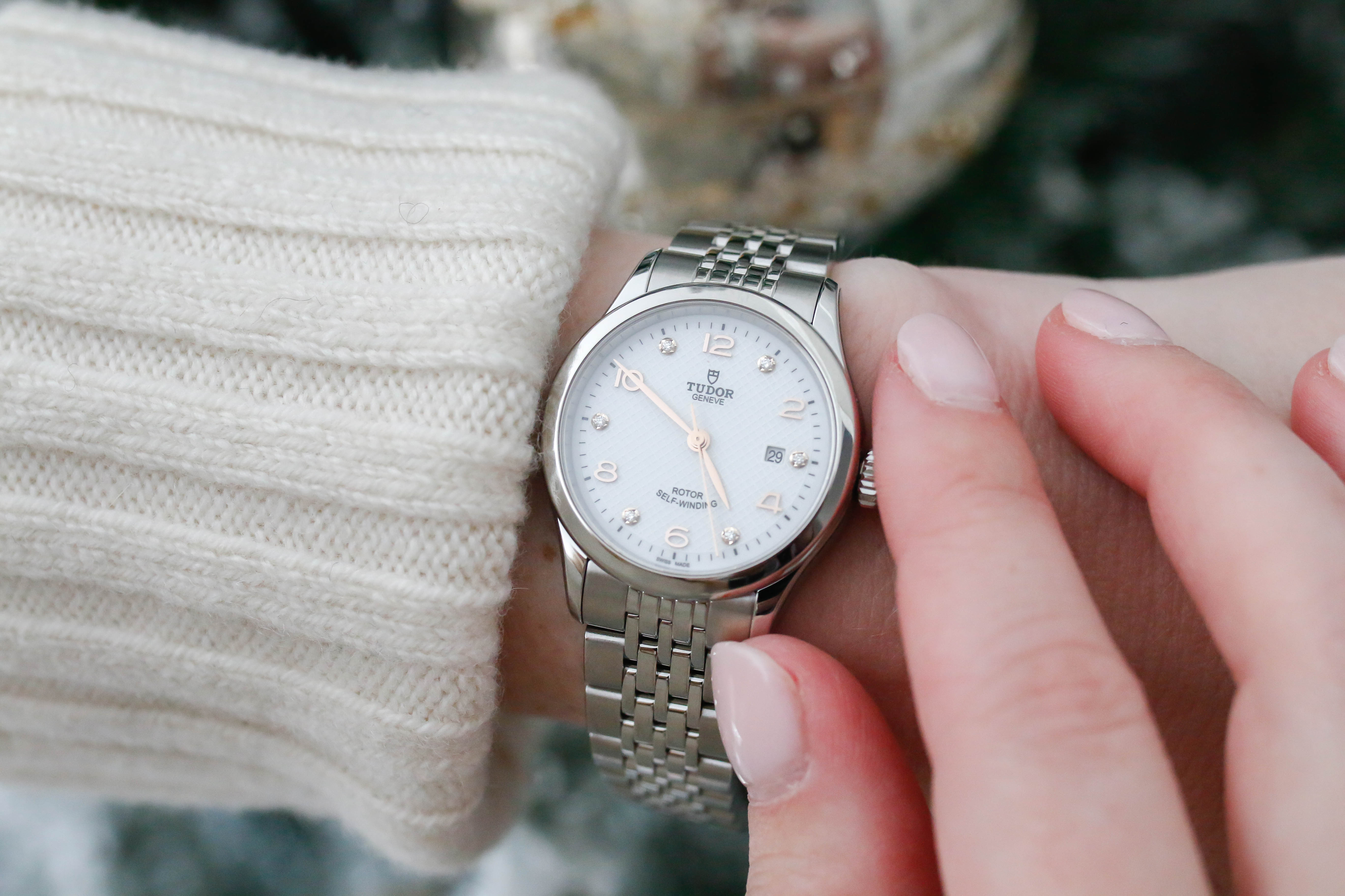 A close-up of a wrist adorned with a Tudor Geneva watch featuring a silver bracelet, a white dial with rose gold accents, diamond hour markers, and a date display, complemented by a cozy white knit sweater.