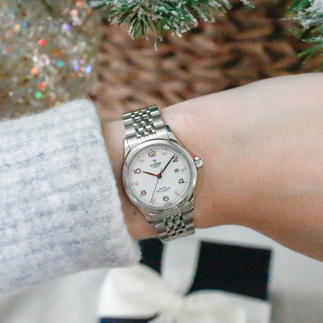 A close-up of a wrist adorned with a Tudor watch featuring a silver bracelet, a white dial with rose gold accents, diamond hour markers, and a date display.